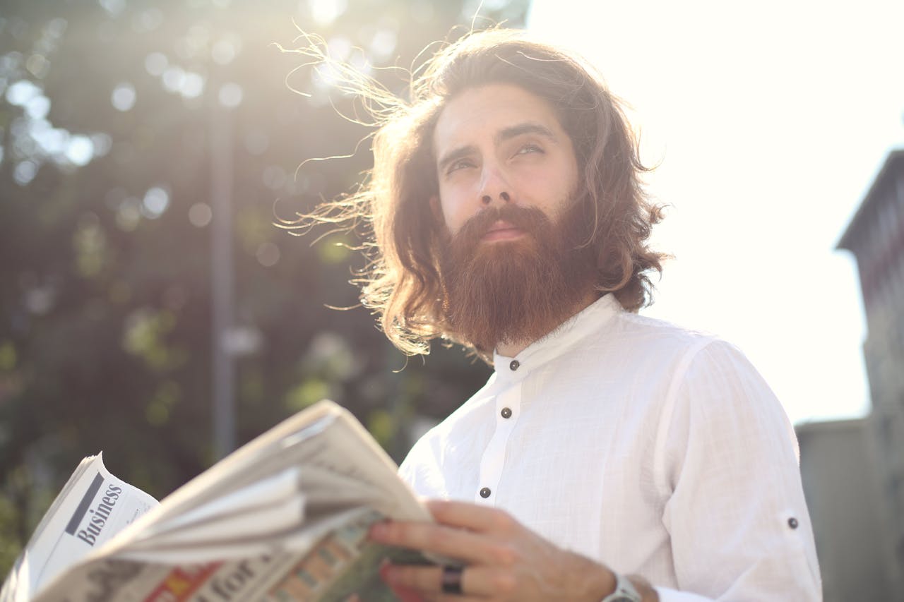services-04 Young bearded man enjoying a sunny day while reading a newspaper outdoors.